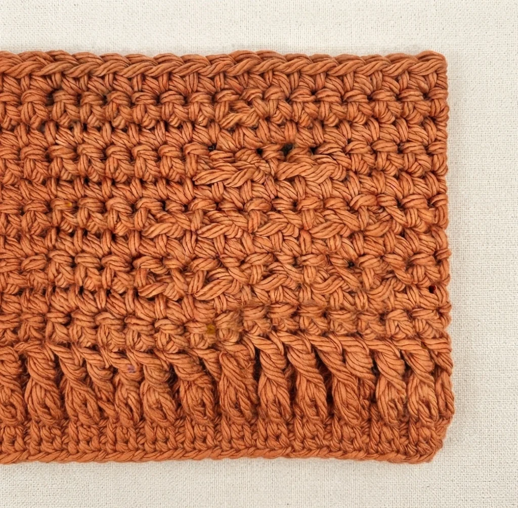 Close-up of rust-orange crochet fabric with dense textured stitches, photographed on a cream fabric background, with the top of a raised pumpkin motif appearing along the bottom edge.