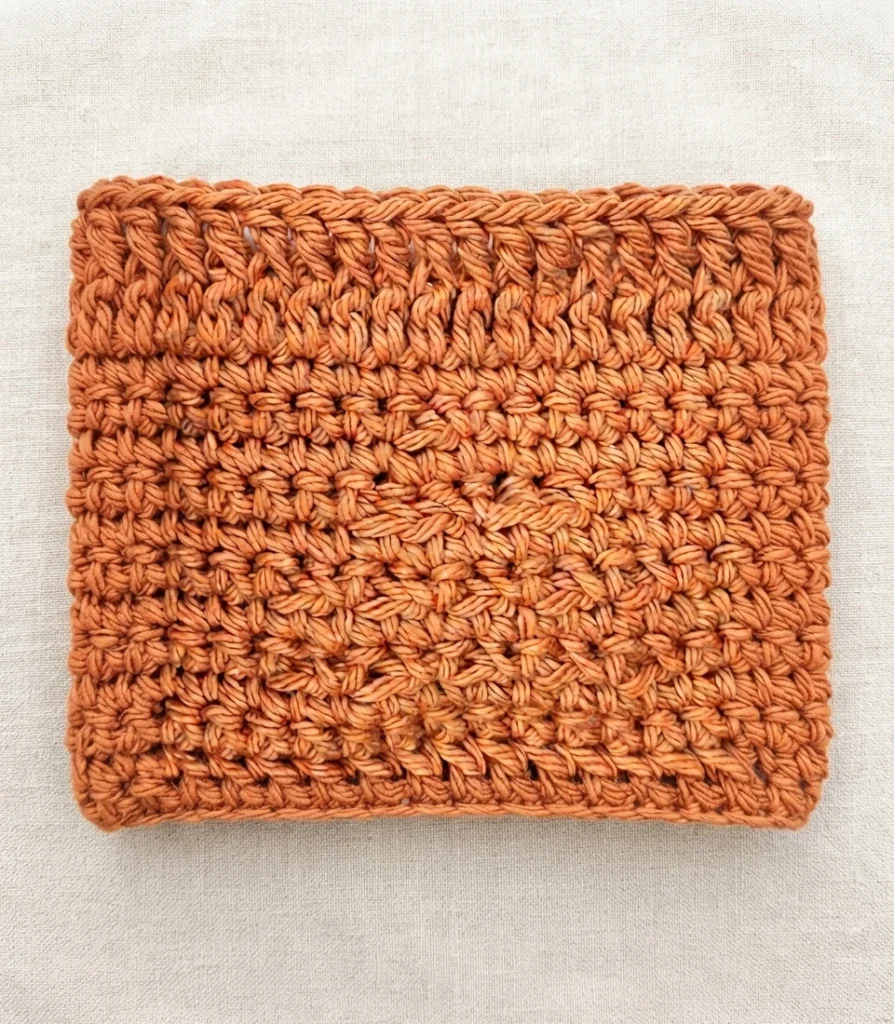 Flat square of orange crocheted cotton fabric with rows of textured post stitches and a subtle raised diamond motif in the center, resting on light beige linen.