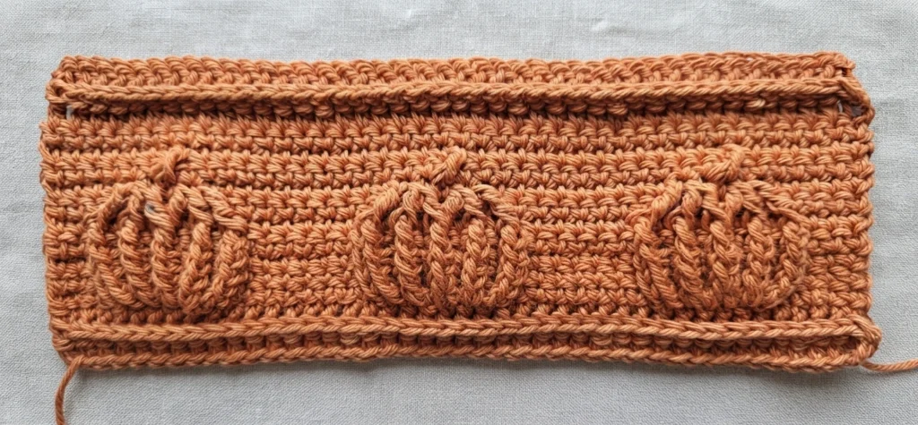 Flat crocheted rectangle in copper orange cotton with three raised pumpkin motifs worked in front post stitches, a ridged top and bottom border, and loose yarn tails at the corners, laid on a grey textured cloth.