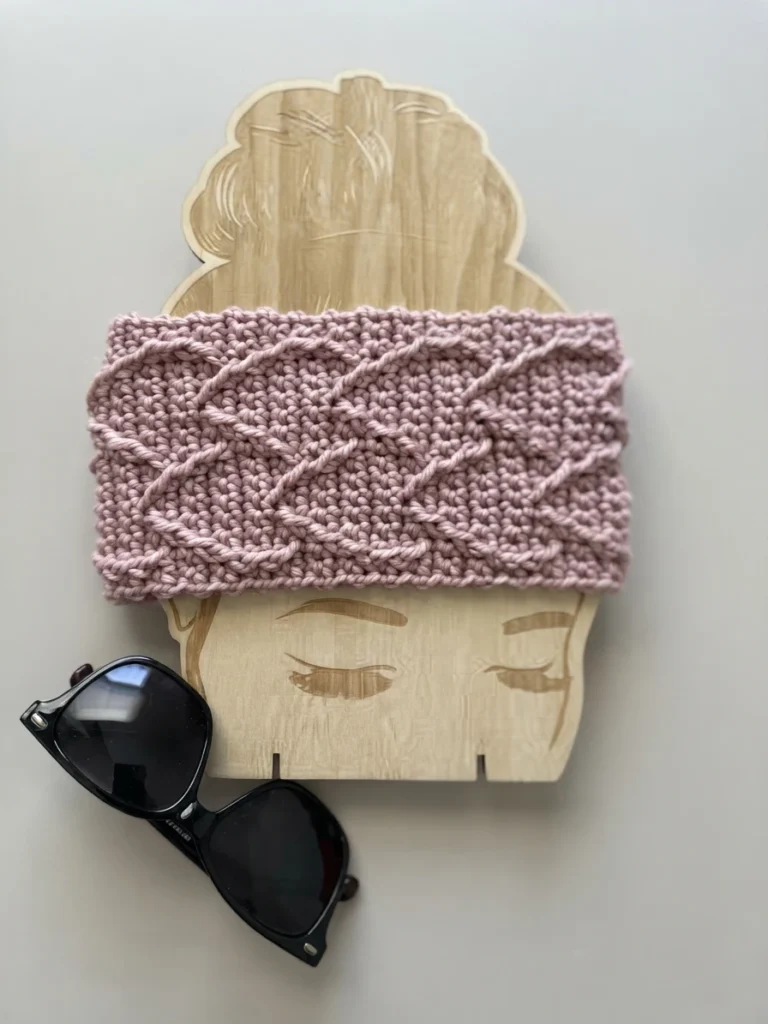 Pink textured crocheted headband with diagonal raised cable-like stitches displayed flat across a wooden head-shaped board, with black sunglasses resting below on a pale background.