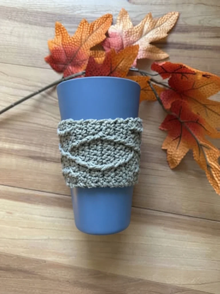 Gray textured crochet cup sleeve with diagonal raised bands around a blue cup, set among artificial autumn leaves on a light wood surface
