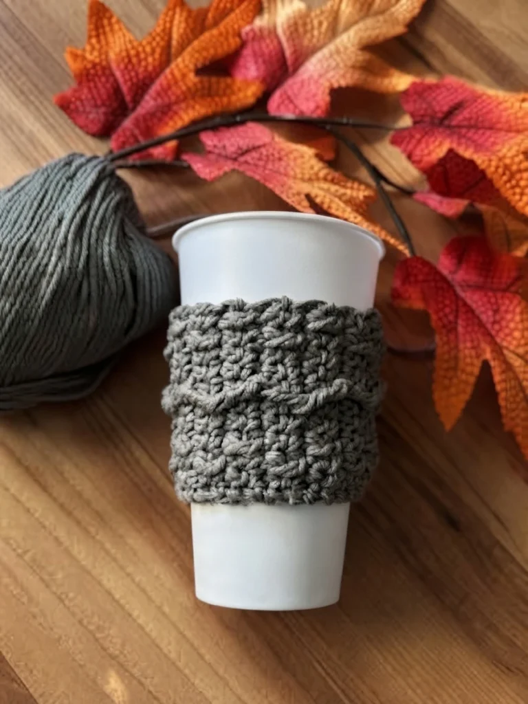 Gray textured crochet cup cozy wrapped around a white paper cup on a wooden table, with a ball of gray yarn on the left and artificial autumn leaves in orange and red behind.