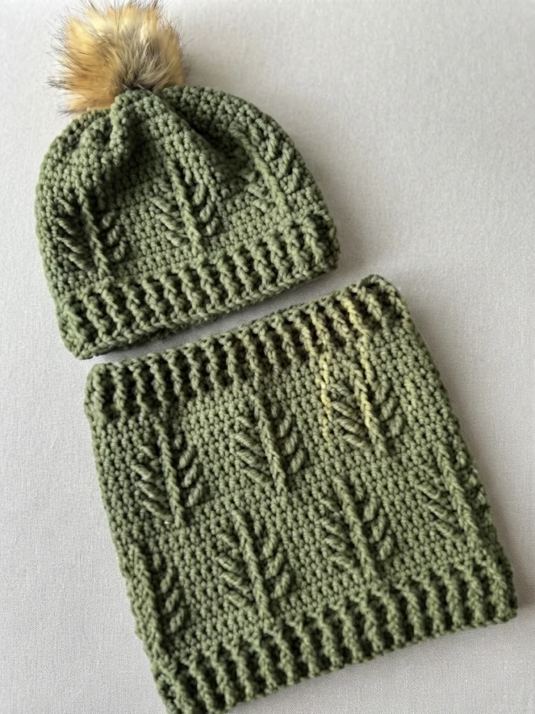 Green crochet hat topped with a faux fur pompom, placed above a matching textured cowl, both laid flat on a light gray surface.