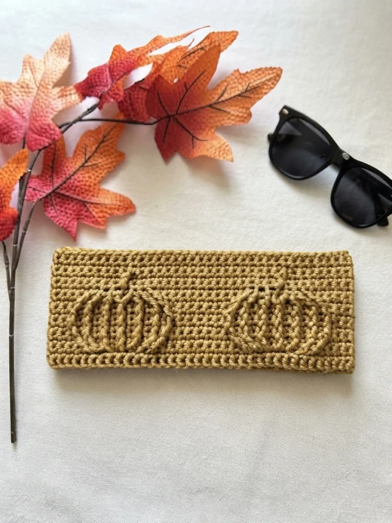 Crocheted tan headband with two textured pumpkin motifs lying flat on off-white fabric, with autumn leaves at the left and black sunglasses at the upper right.