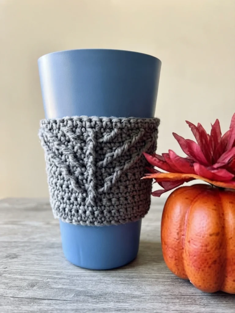 Grey crocheted cup cozy with a raised leaf motif wrapped around a blue tumbler, next to an orange pumpkin and a red artificial flower on a grey wooden table.