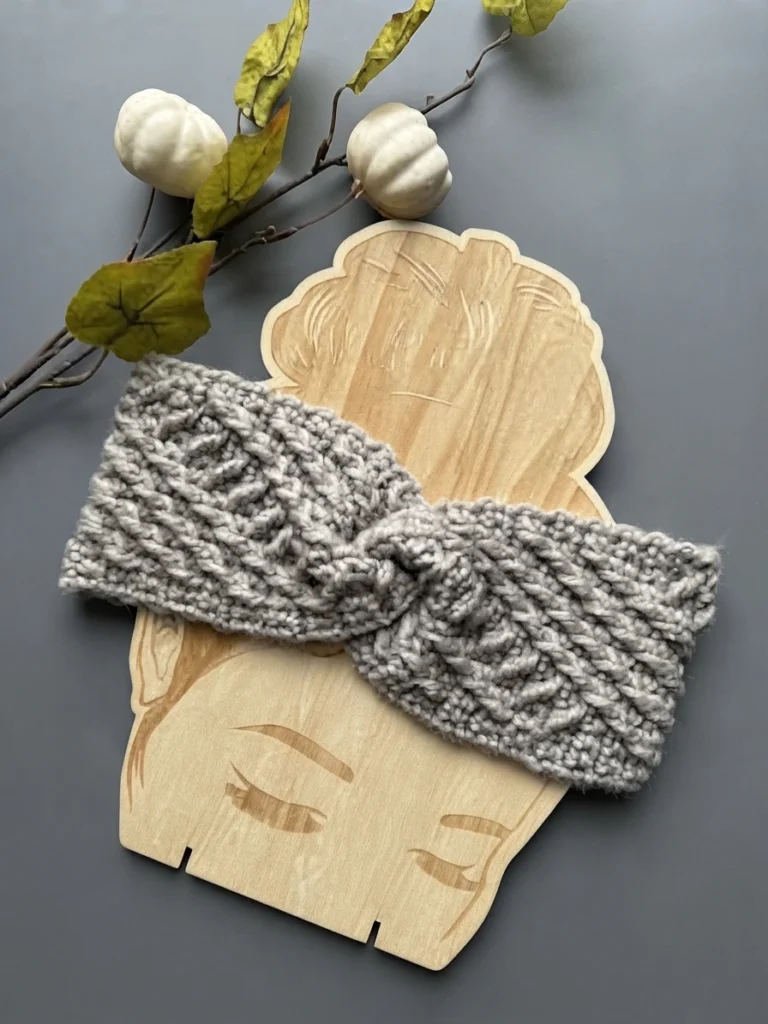 Gray textured headband with a twisted front displayed on a carved wooden head-shaped form, surrounded by small white pumpkins, green leaves and thin branches on a plain gray background.
