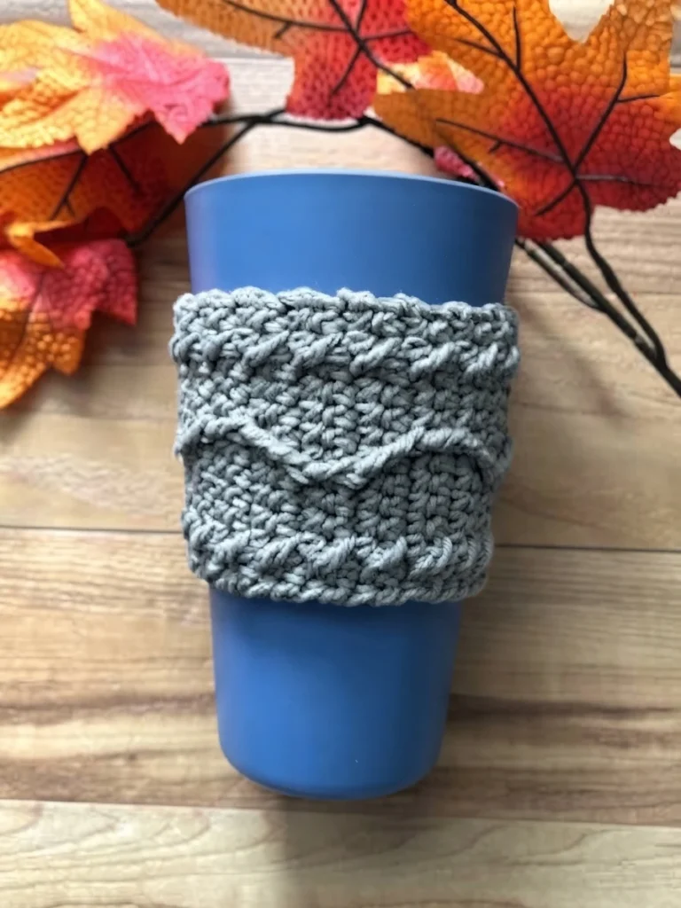Gray textured crochet cup cozy with wavy raised lines wrapped around a blue cup on a wood surface, with artificial orange and red autumn leaves behind it.