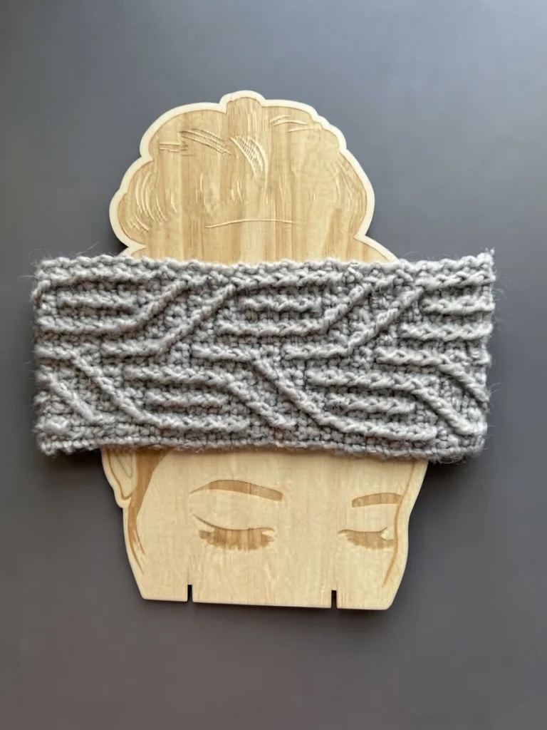 Gray crocheted headband with raised wavy lines displayed on a light wooden head-shaped form against a plain gray background