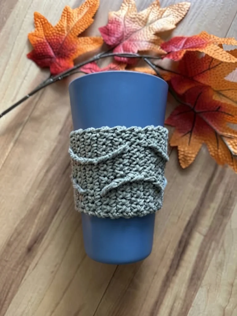 Grey crocheted cup cozy with wavy textured ridges wrapped around a blue tumbler, set on a wood surface with artificial autumn leaves in orange, red and cream.