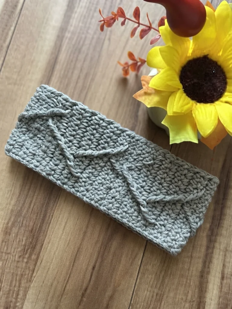 Gray crocheted headband with a raised cable pattern lying flat on a wooden floor beside a yellow sunflower and a sprig of orange leaves