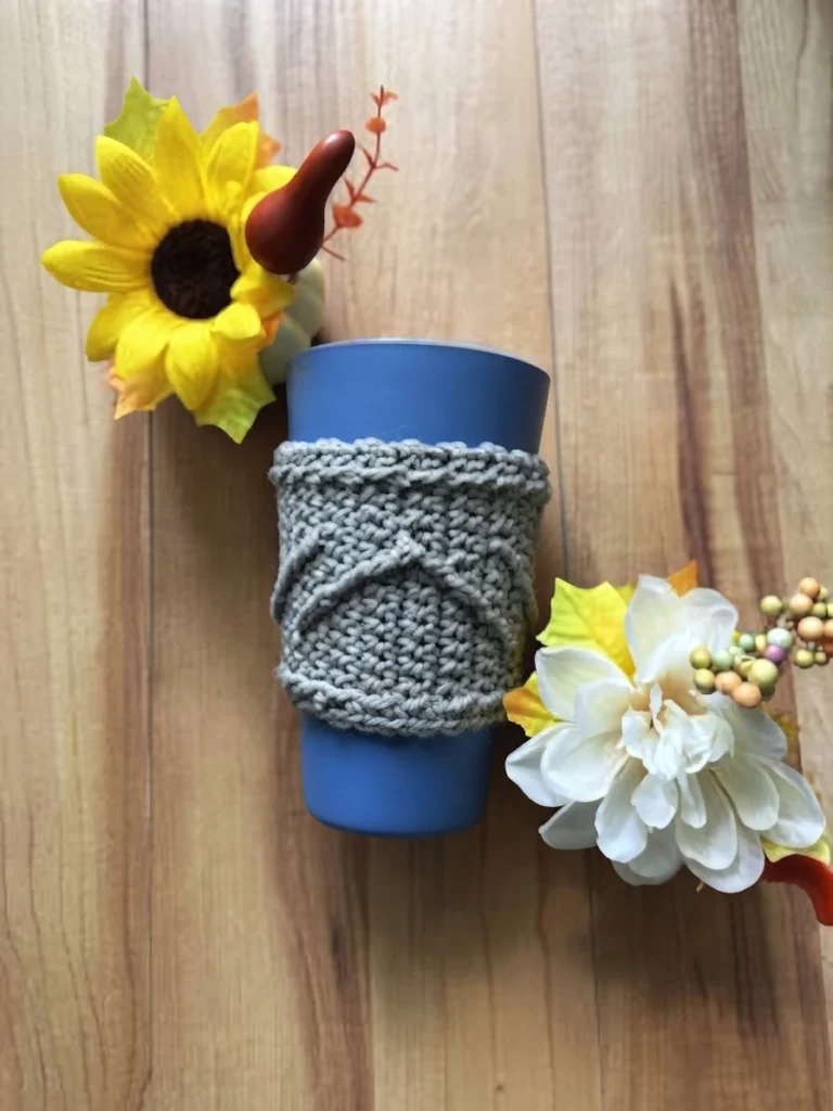 Light grey crocheted cup sleeve with raised diagonal ridges wrapped around a blue cup on a wooden surface, with a yellow sunflower to the left and a white flower with berry sprigs to the right.