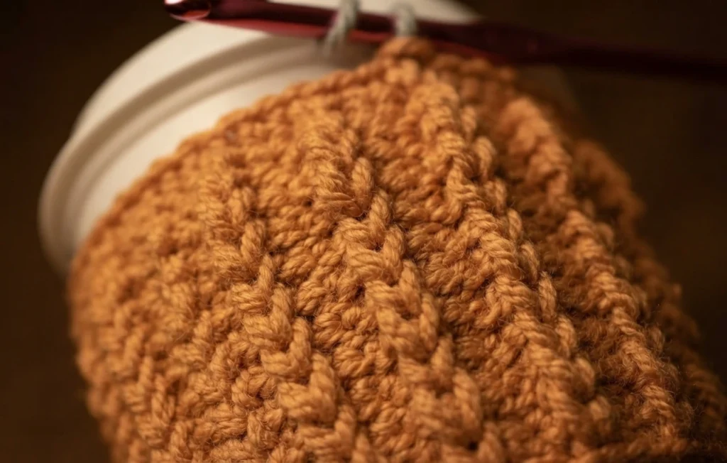 Close-up of orange crochet fabric worked in textured post stitches, wrapped around a white cup, with a red crochet hook still resting in the top edge of the work.