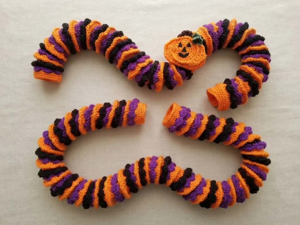 Two crocheted spiral garlands in orange, purple and black laid flat on white fabric, one with a small crocheted jack-o-lantern pumpkin attached