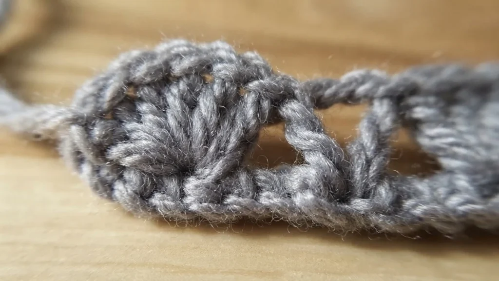 Close-up of light gray crocheted yarn on a wooden surface, showing a dense cluster of stitches beside an open chain space.