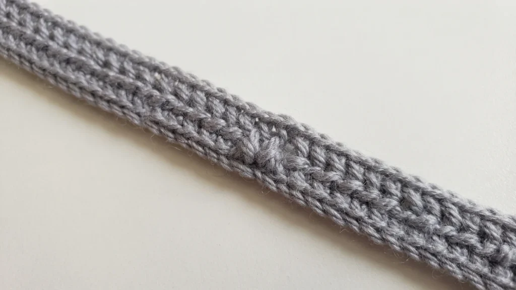 Close-up of a gray crocheted strip worked in rows of tall stitches, lying diagonally on a plain light background