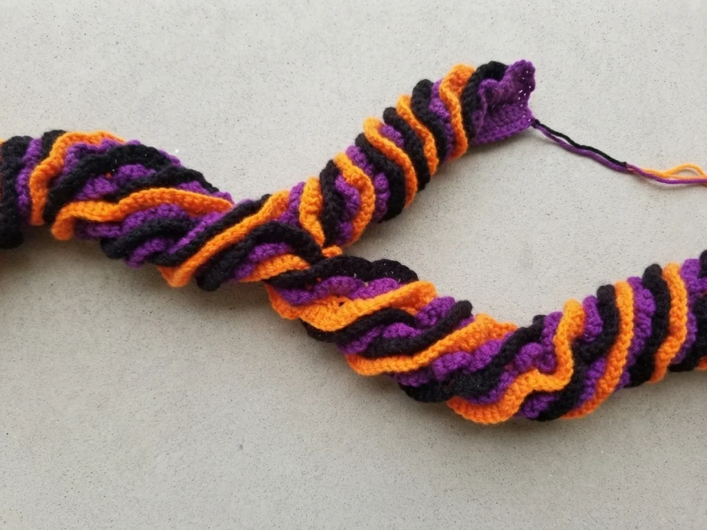 Three crocheted garlands in black, purple and orange twisted together and lying on a pale flat surface, with loose yarn ends visible at one end.