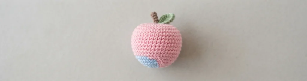 Pomme en crochet rose garnie, avec une petite zone bleue à la base, une tige marron et une feuille verte, posée sur un fond gris clair uni.