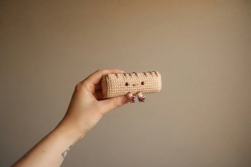 A hand holding a small beige crocheted rectangular plush with a stitched face and short legs tipped with purple beads, photographed against a plain beige wall.