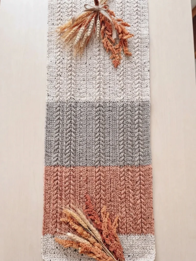 Long crochet table runner with cream, gray, and rust-colored sections and raised stitch patterns, decorated with dried foliage.