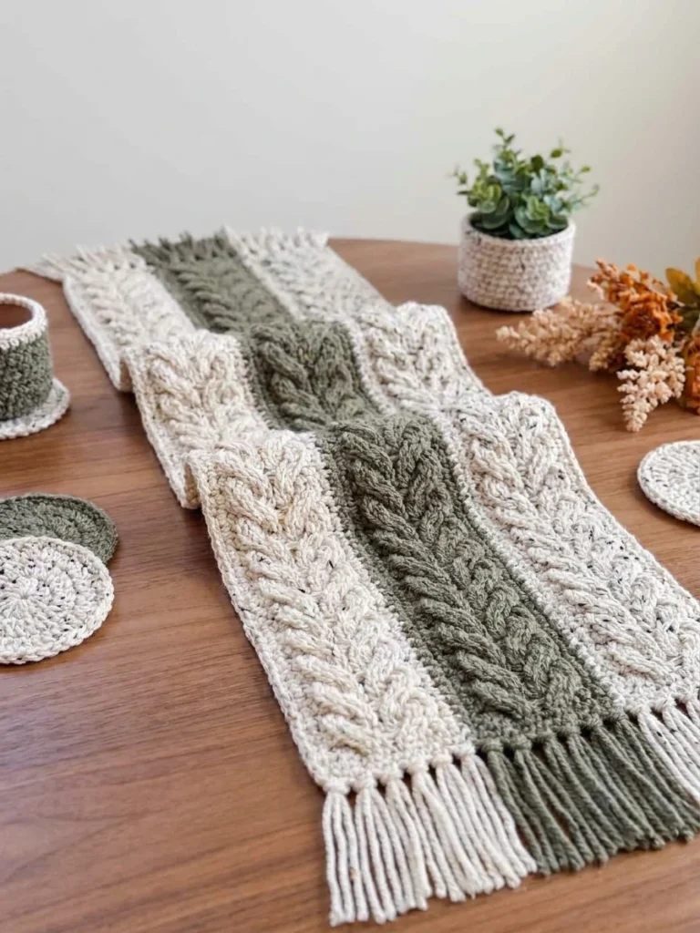 Chemin de table crocheté à bandes crème et vert sauge, avec des franges, posé sur une table en bois entourée de petits accessoires décoratifs.