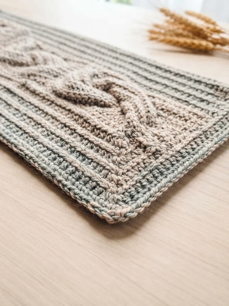 A beige and gray crocheted table runner with a raised central motif and textured border, photographed on a wooden surface beside dried wheat.
