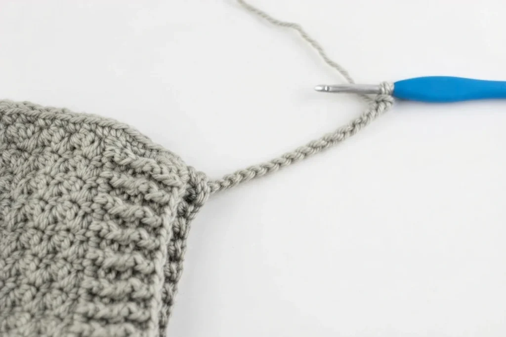 Grey crochet bonnet in progress with a chain for a tie extending to a blue handled crochet hook on a white background