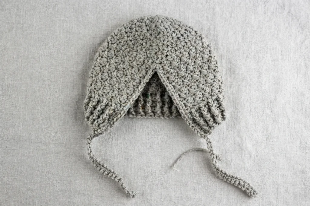 Gray crochet baby bonnet without ears laid flat, showing a V opening, ribbed brim and two crocheted ties with a loose yarn end