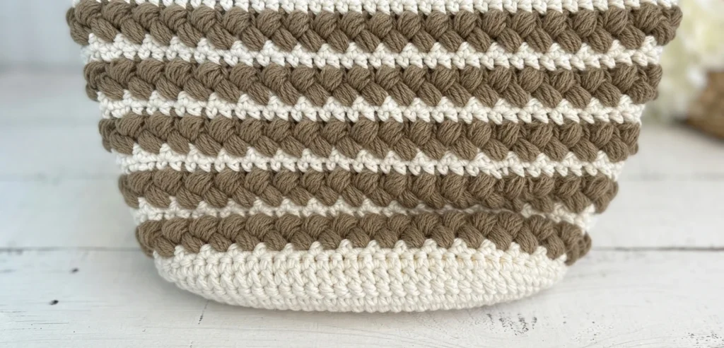 Close-up of a crocheted tote bag body in white and taupe showing alternating rows of braided puff stitches with a visible slanted seam.