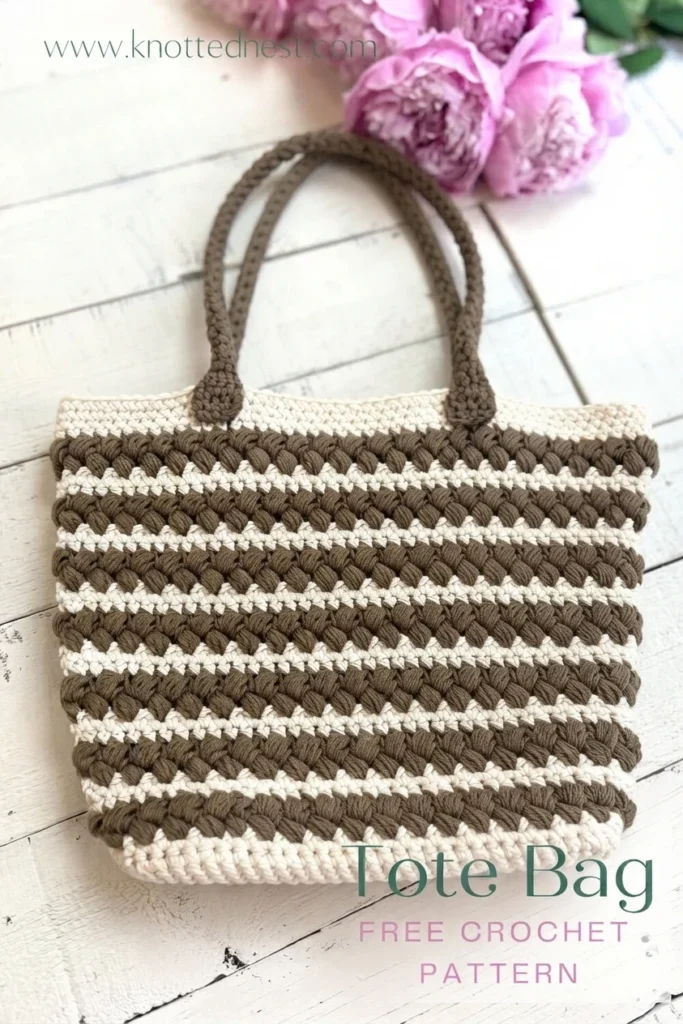 Crochet tote bag with taupe braided stripes and cream rows, finished with crocheted handles, photographed on a white wooden surface with pink flowers and a free pattern text overlay.