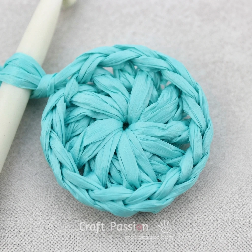 Close-up of a turquoise raffia crochet circle with a white crochet hook on light fabric.