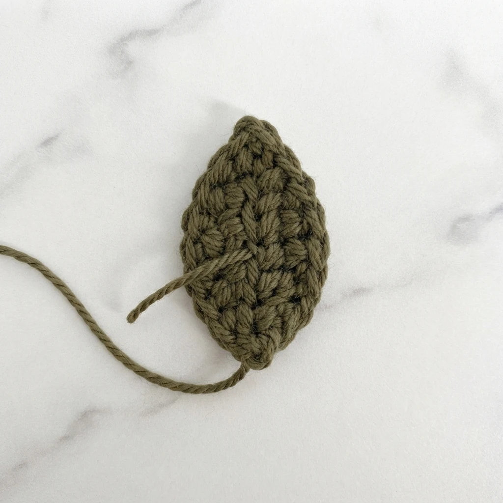 Olive green crocheted leaf with a loose yarn tail on a white marble surface.