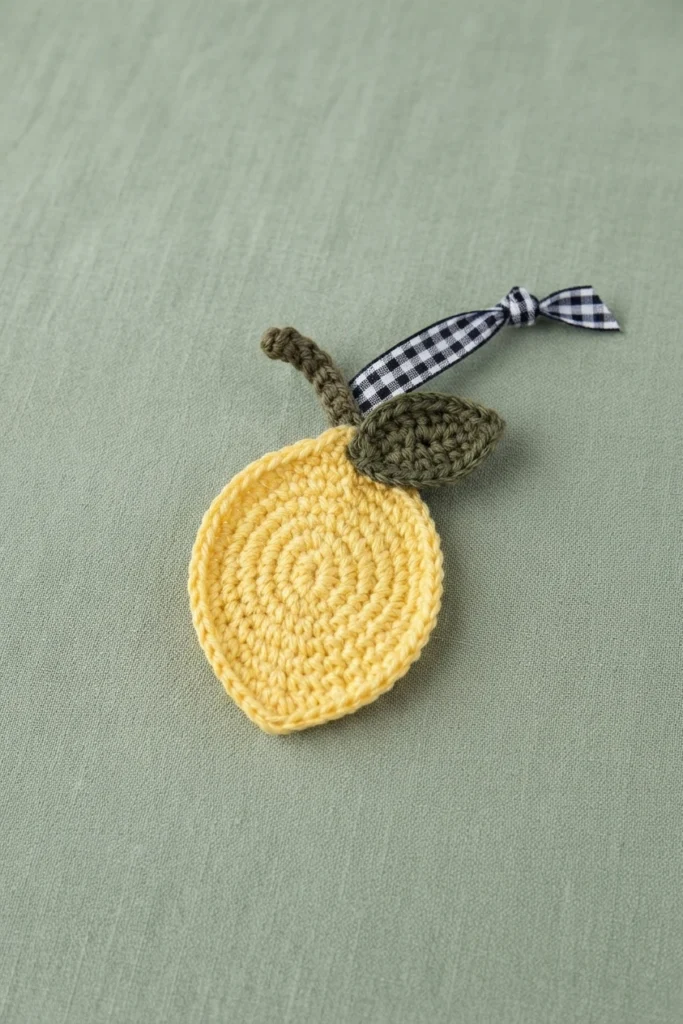 Crocheted yellow lemon motif with olive green leaf and stem beside a black-and-white gingham ribbon, lying on sage green fabric.