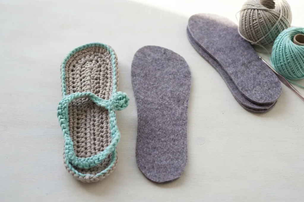 One crochet flip-flop with a taupe sole and mint straps beside grey felt soles, yarn balls and a crochet hook on a light surface