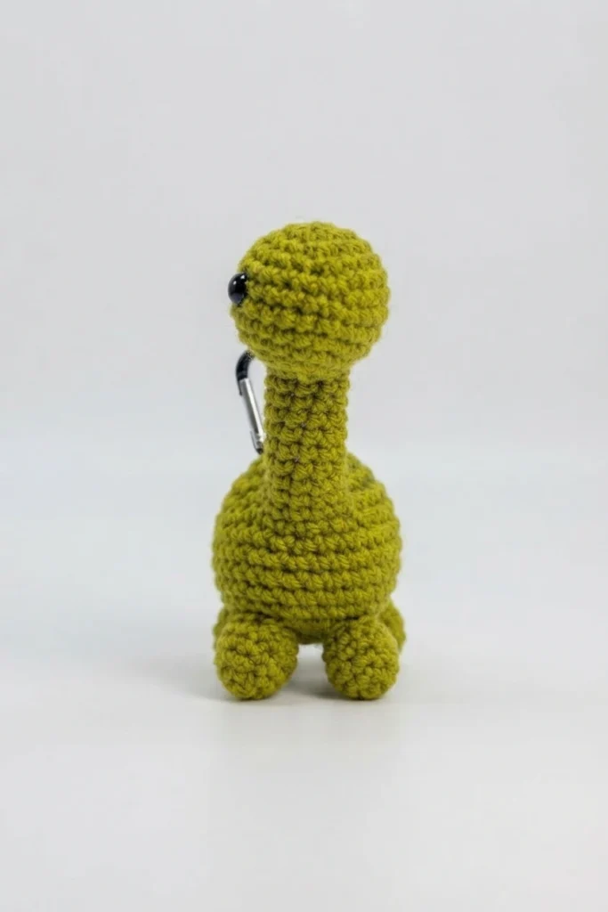 Small green crochet brontosaurus with a long neck, black safety eye and a small carabiner clip on its back, standing against a plain light background.