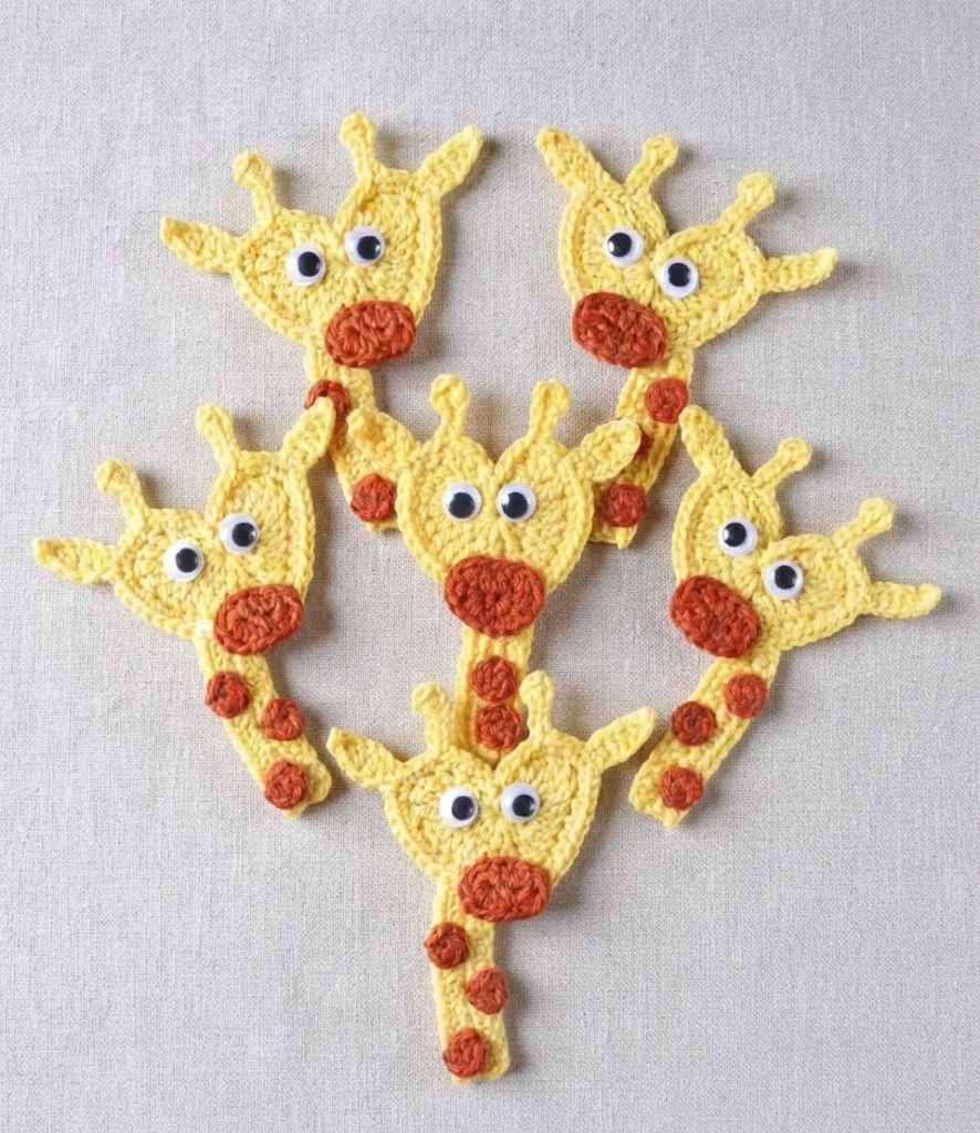 Six yellow crocheted giraffe appliques with rust colored muzzles and spots and googly eyes arranged on light textured fabric.
