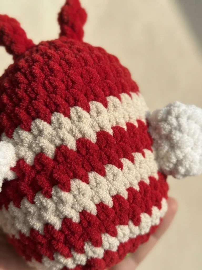 Close-up of a crocheted bee body worked in red and white zigzag stripes, with two small red antennae on top and a fluffy white wing at the side, held in a hand against a soft pale background.
