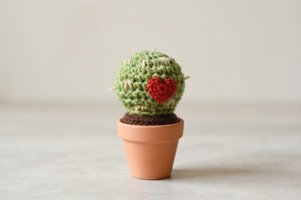 A green crocheted round cactus with light spines and a red heart on the front, in a small terracotta pot with brown soil
