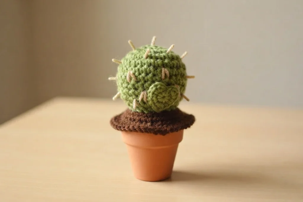 A single green crocheted cactus with tan spines and a green heart on the front, in a terracotta pot on a light wooden table