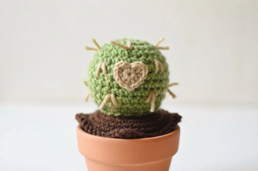 A single round green crocheted cactus with tan spines and a beige heart on the front, in a terracotta pot with brown crocheted soil