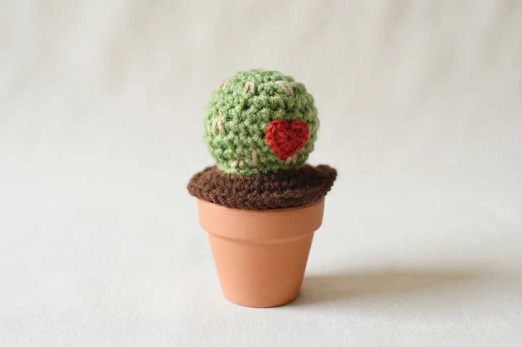 A round green crocheted cactus with small tan spines and a red heart on its front, in a terracotta pot with brown crocheted soil
