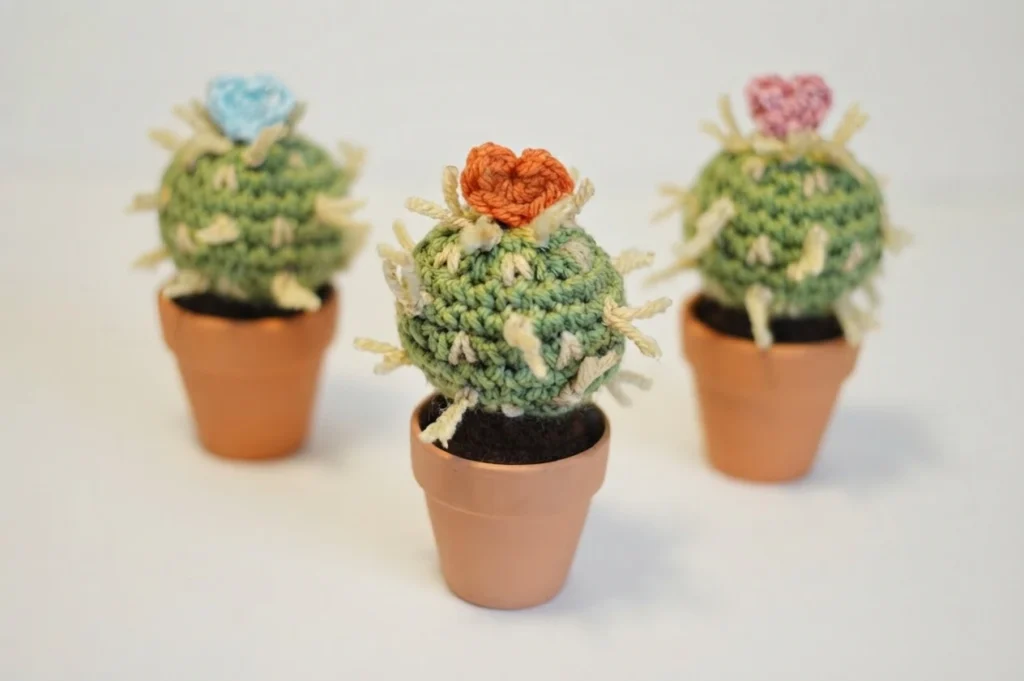 Three small green crocheted cacti in terracotta pots with cream spines, topped with blue, orange and pink crocheted hearts