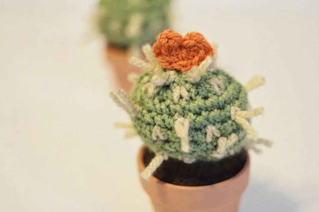 Close-up of a green crocheted cactus with cream spines and an orange heart on top in a terracotta pot, with another cactus blurred behind it