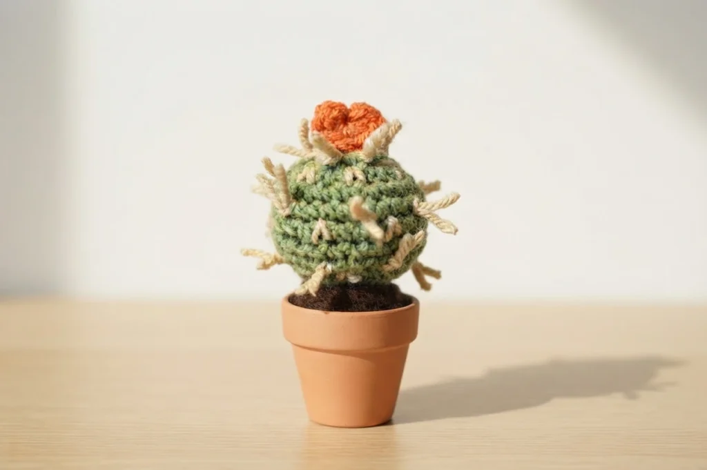 A crocheted green round cactus with cream yarn spines and an orange heart on top, in a terracotta pot on a wooden surface