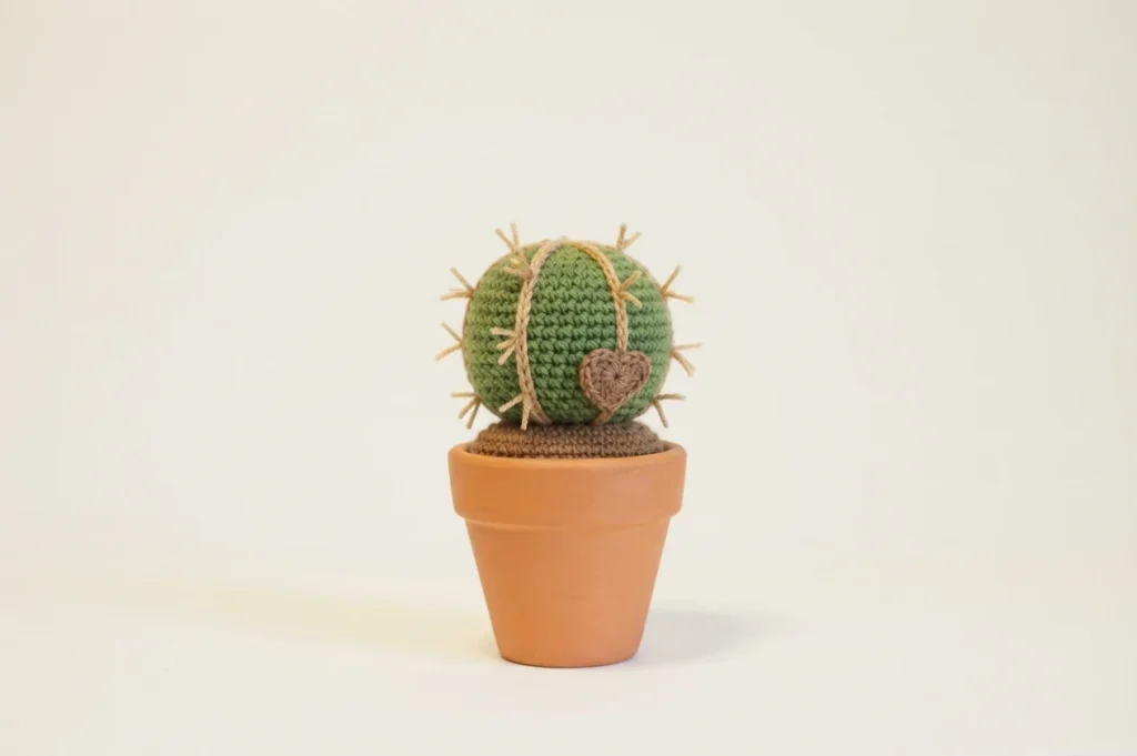A single green crocheted cactus with tan spines and vertical cords and a brown heart on its front, sitting in a terracotta pot with brown crocheted soil on a plain light background