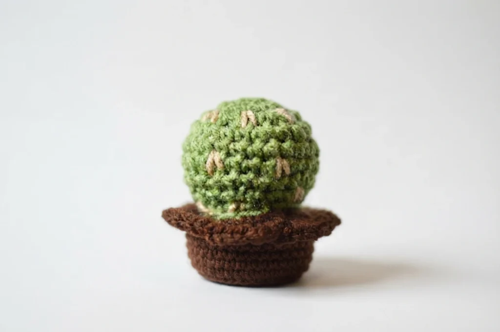 A single round green crocheted cactus with small tan V shaped stitches, sitting in a brown crocheted pot on a white background