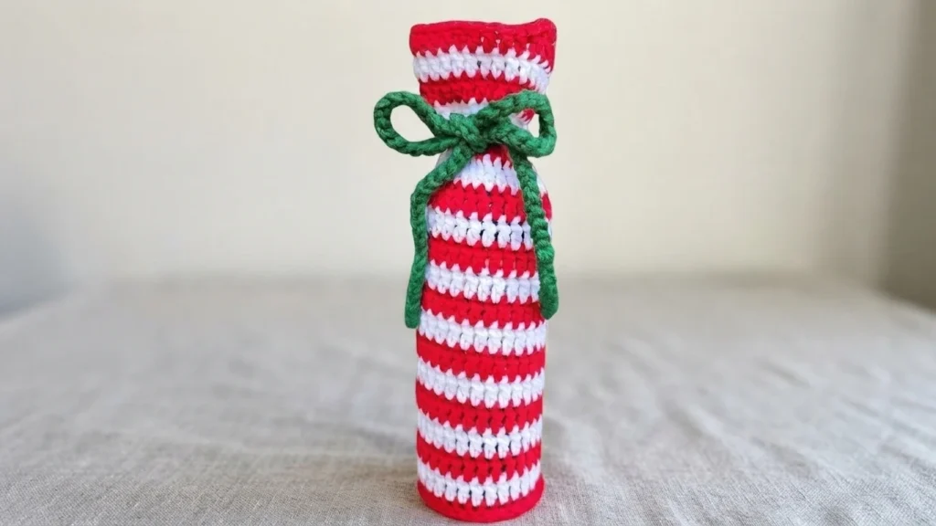 Crocheted wine bottle bag in red and white candy cane stripes with a green tied bow, standing upright on a light linen surface against a plain wall
