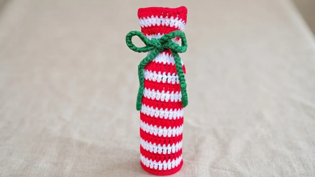 Finished red and white striped crochet wine bottle bag tied at the neck with a green yarn bow, shown on a plain beige linen surface