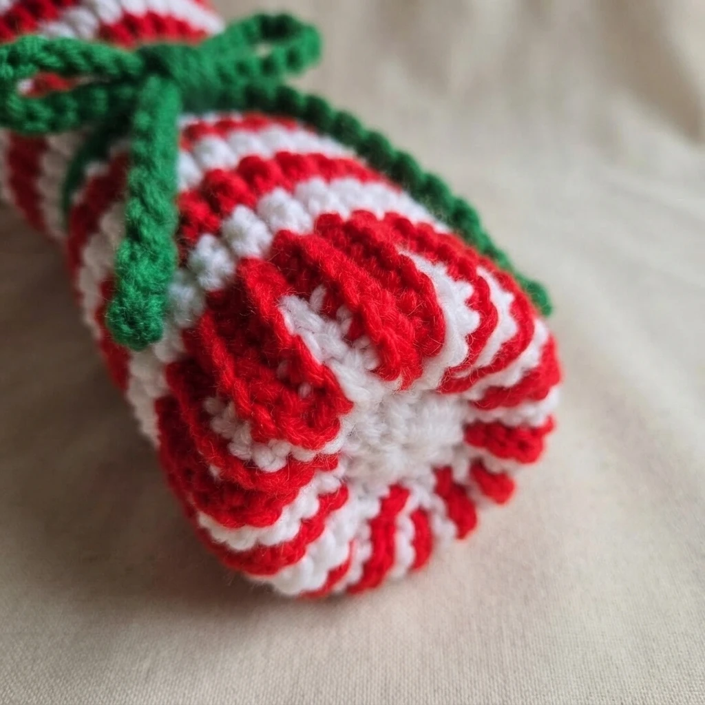 Crochet wine bottle bag in red and white candy cane stripes with a green tied bow, lying on its side on a plain cream cloth so the round bottom edge faces the camera