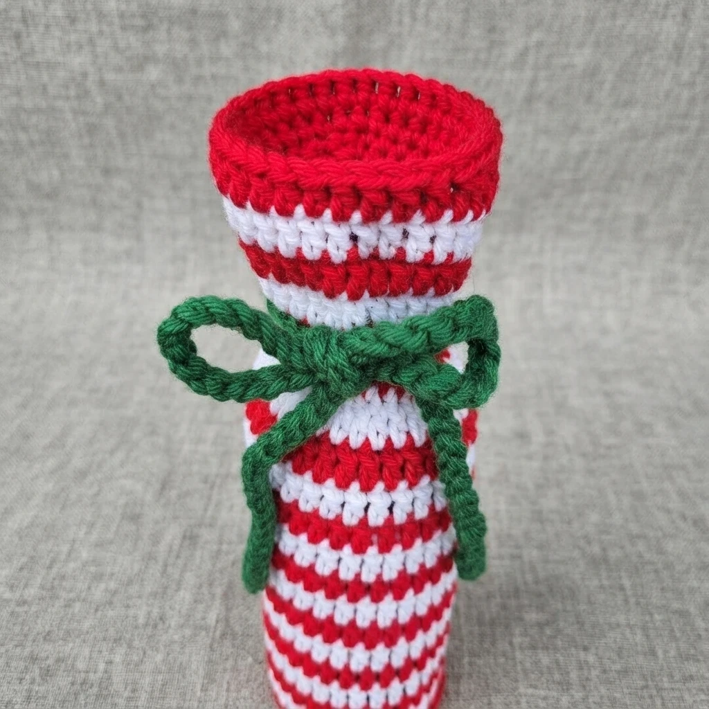 Five identical crochet wine bottle gift bags in red and white candy cane stripes, each tied with a green yarn bow, lined up on a wooden plank against a blurred field background.