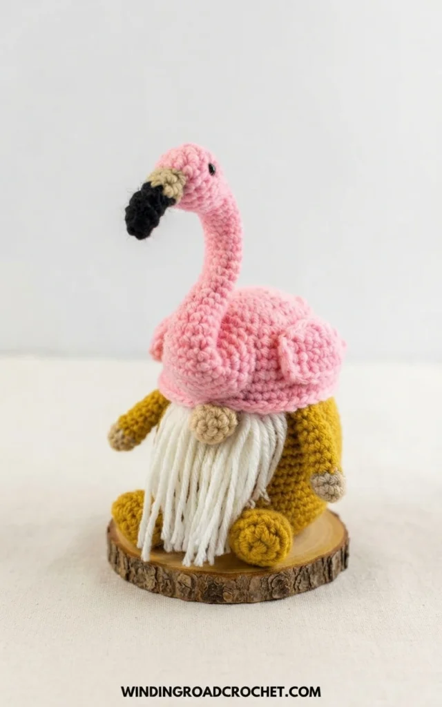 Crocheted flamingo gnome with a pink flamingo hat, black and beige beak, long white yarn beard and mustard yellow body, seated on a wood slice against a plain cream background.